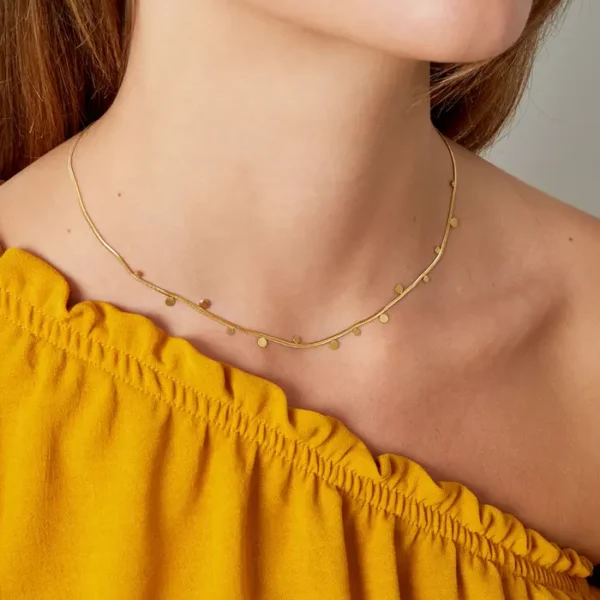 Vrouw draagt gouden ketting en gele top.